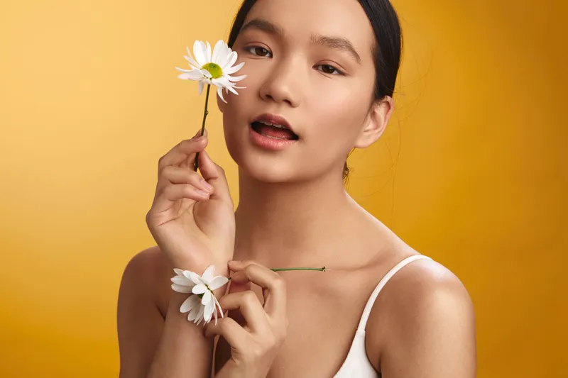 asian woman holds a white flower near her face against a warm yellow backdrop, a soft smile and serene gaze conveying gentle elegance and creative expressiveness.