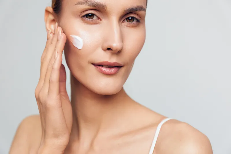 A young woman with clear skin applies white cream to her cheek in a close-up portrait on a light background. Concept for skincare, beauty routine, and healthy skin care.