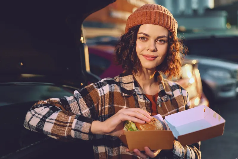 Woman with burger and takeaway box by open car trunk on urban street in evening, casual smile and plaid jacket; candid lifestyle, authenticity, golden hour glow, mindful living and emotional