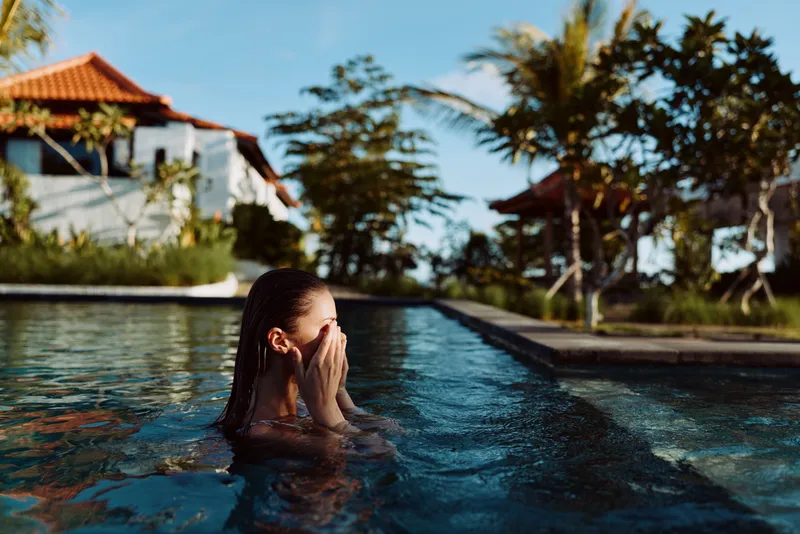Young woman with wet hair covering her face while standing in an outdoor pool. Tropical trees and houses in the background. Concept of relaxation and lifestyle.