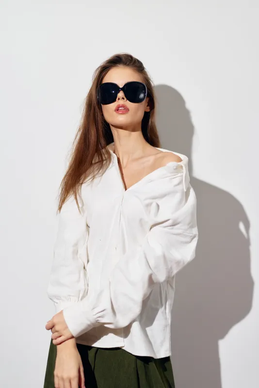 Модная модель демонстрирует трендовую oversize-белую рубашку в сочетании с эффектными солнцезащитными очками, стоя на минималистичном белом фоне, который подчёркивает её стильный образ