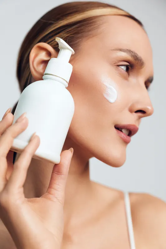 Young woman applying white cream on cheek holding plastic pump bottle close to face, skincare routine, clean skin, beauty treatment, soft lighting, minimal background