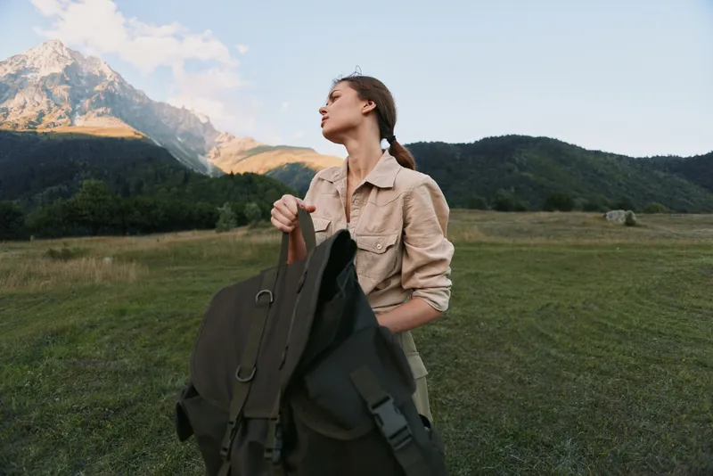 Woman dressed in casual jacket stands in an open field with a backpack, mountains in the background, showcasing outdoor lifestyle and travel moments, calm and adventurous atmosphere.