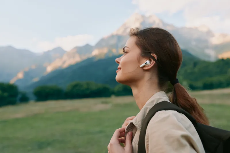 Outdoor portrait of a young woman with a backpack listening to music in a green field with distant mountains under a clear sky