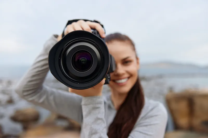 Young woman smiling while holding a camera outdoors by the seaside, showcasing a passion for photography and a connection with nature