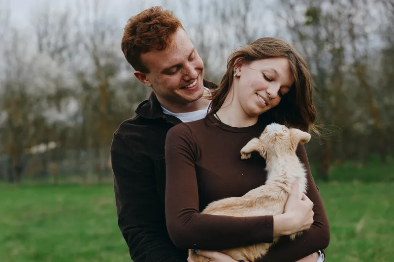 Couple smiles as they cradle a goat kid in a sunny field, showing affection and bonding. Outdoor lifestyle scene with natural light, grass, and trees, conveying warmth and happiness.