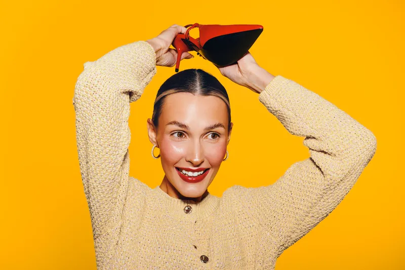 Smiling woman in beige sweater pours red wine from a bottle above her head on bright yellow background. Concept of fun, celebration, and joyful lifestyle with vibrant colors.