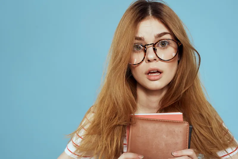 Энергичная студентка с блокнотами и в очках позирует на синем фоне