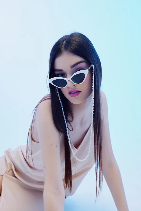 Fashionable woman with long dark hair wearing white sunglasses and a beige dress poses against a light blue background. Concept of beauty, fashion, and style.