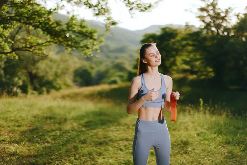 Happy young woman exercising outdoors with resistance bands, dressed in gray sportswear, enjoying fitness in a lush green park surrounded by nature