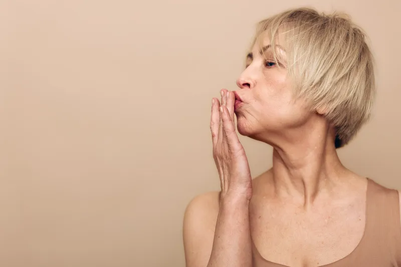 Portrait of a mature woman with short blonde hair blowing a kiss, isolated on beige background. Concept for affection, love, emotion, and positive human connection with soft lighting.