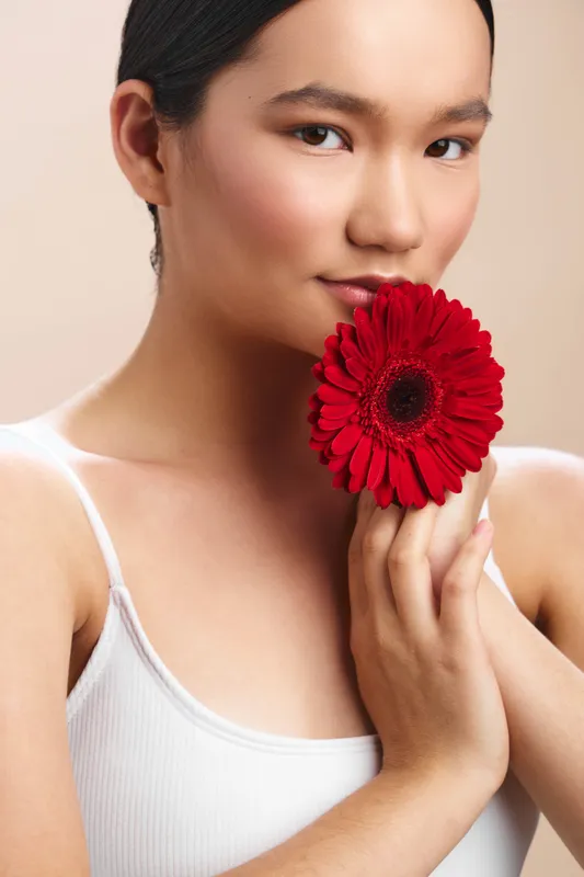 Asian woman softly smiles while holding a red flower near her face, posing in a simple studio with gentle lighting, conveying elegance, calmness, and natural beauty.