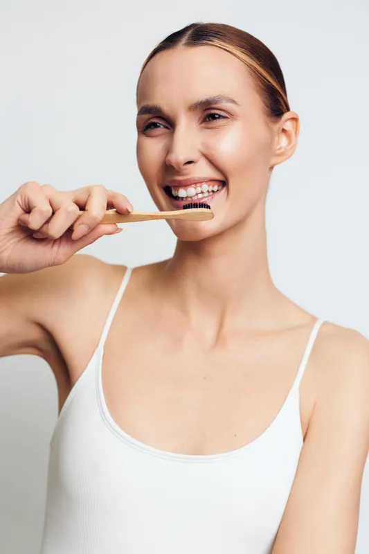 Young woman brushing teeth with natural bamboo toothbrush smiling gently white background casual tank top healthy dental care clean hygiene routine