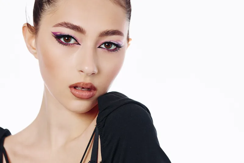 Close-up of a young woman with creative purple eyeliner and glossy nude lips wearing a black top. Studio portrait on a white background focused on fashion and beauty trends.