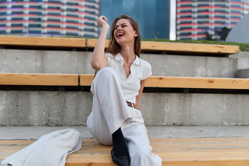 Business woman in a stylish outfit laughing joyfully on outdoor steps with a modern city background, showcasing confidence and professionalism.