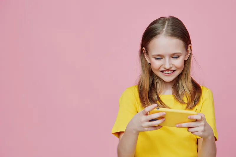 Happy girl in a yellow shirt smiles while using a smartphone against a pink background, showcasing joy and technology in a vibrant, playful setting