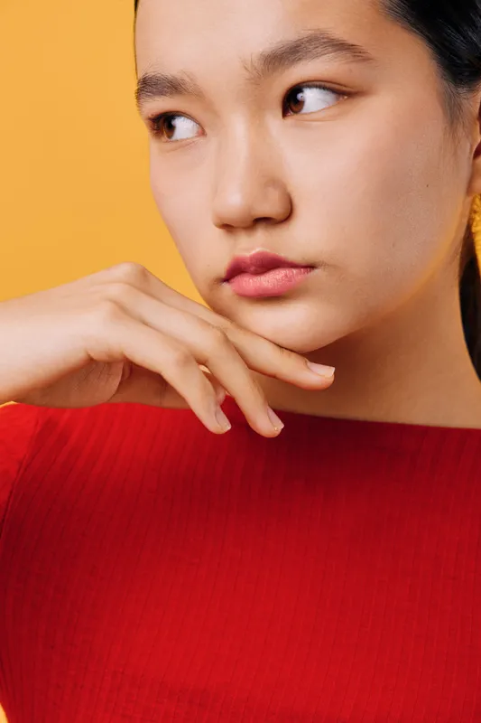 Young woman with smooth skin in red top poses looking aside with hand near face on yellow background. Closeup portrait of thoughtful female model with natural makeup and clear complexion.