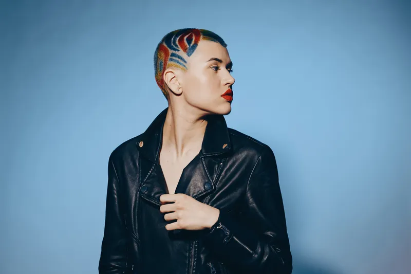 woman with colorful shaved hair and leather jacket posing sideways with bold red lipstick against blue background, showcasing modern edgy style and confident attitude.