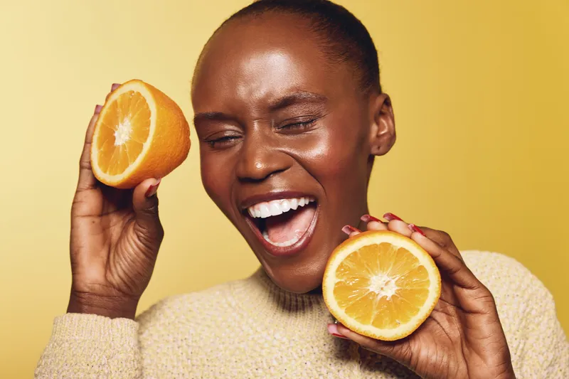 Happy African woman with dark skin holding two halves of fresh orange near face, smiling with closed eyes on yellow background. Concept of healthy lifestyle, vitamin, and natural beauty.