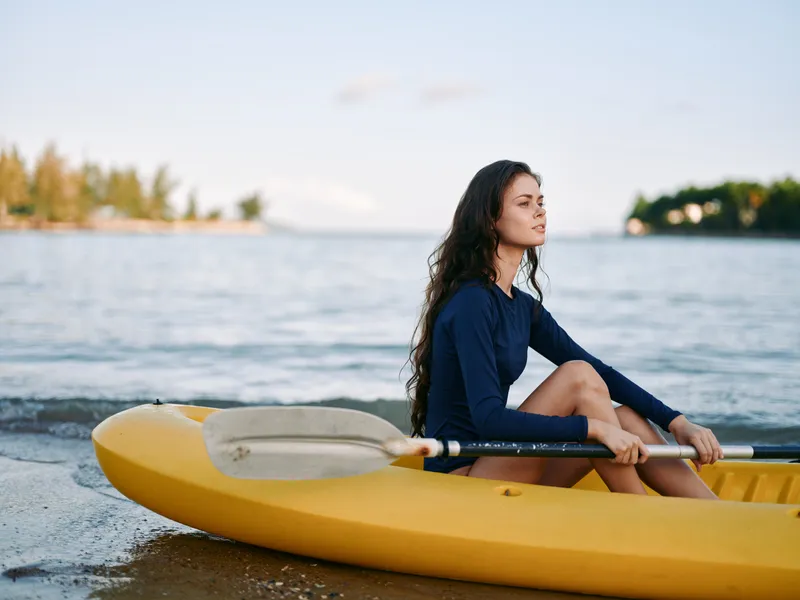 A young woman sits in a yellow kayak on a calm shoreline, gazing toward the horizon as warm light bathes the water and distant trees.