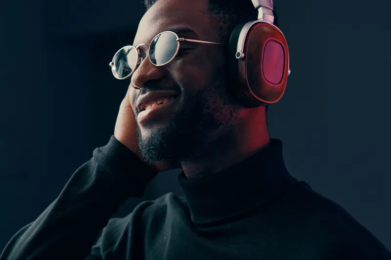 A joyful young Black man wearing stylish headphones and sunglasses, enjoying music in a modern setting with a dark blue background that enhances the mood