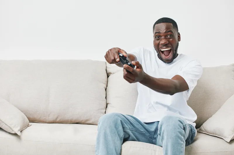 Excited young African man playing video games on a cozy couch, expressing joy and enthusiasm while wearing a casual white t shirt and jeans
