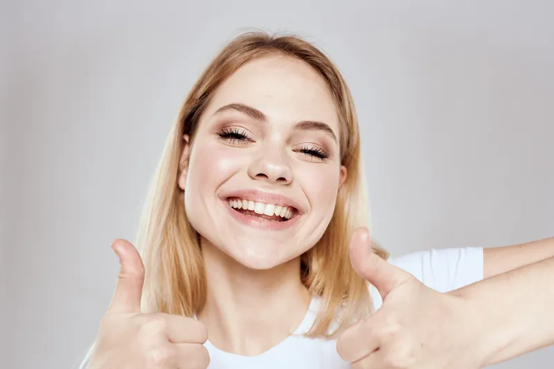 Весёлая блондинка в белой футболке жестикулирует руками на светлом фоне