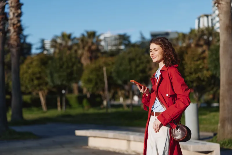 Young woman with curly hair in a red coat walks outdoors, smiling while checking her phone in a sunny park setting, with palm trees and a bright urban background