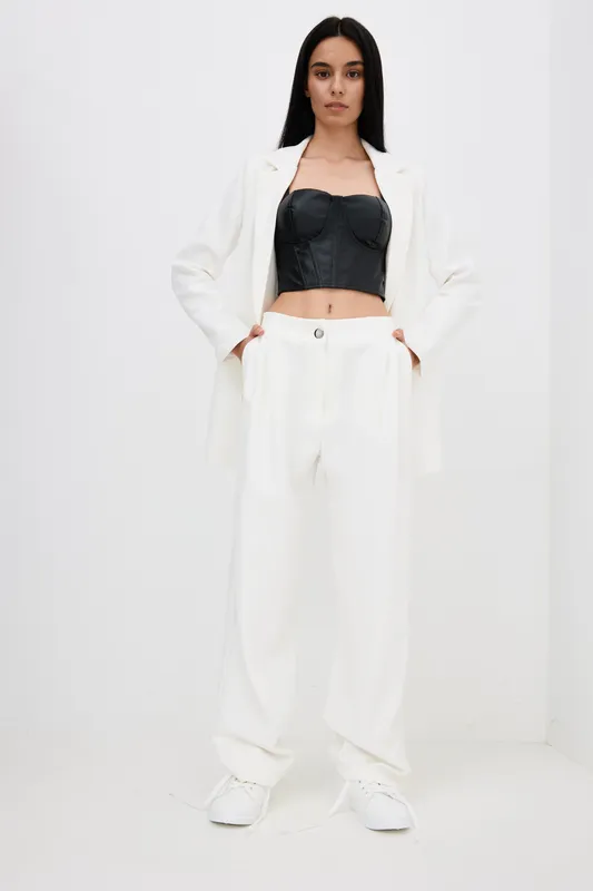 Young woman wearing white pantsuit and black corset top stands confidently against plain white background, showcasing modern fashion style and casual elegance.