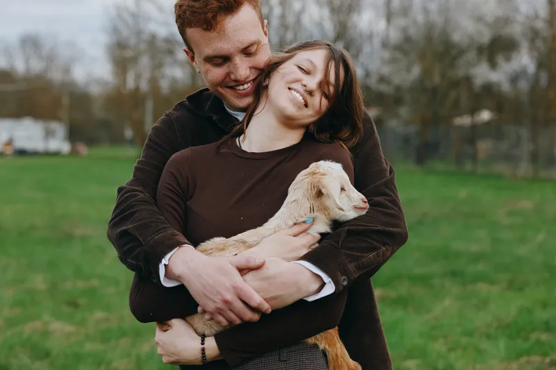 Joyful couple outdoors embracing a goat kid, warm natural light on a rural field, showcasing lifestyle, affection, and animal companionship in a relaxed candid mood.