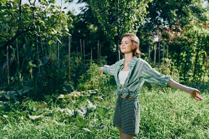 Young woman stands in a sunlit garden with arms outstretched, wearing striped shorts and a green cardigan, expressing freedom and joy.