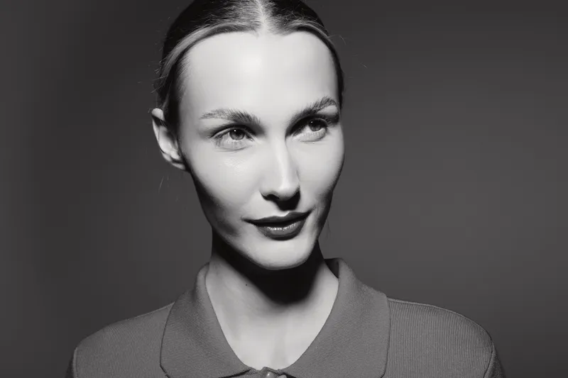Close-up portrait of a fair skinned Caucasian woman with slicked back hair, wearing a textured top, looking sideways with a subtle smile on a dark studio background.