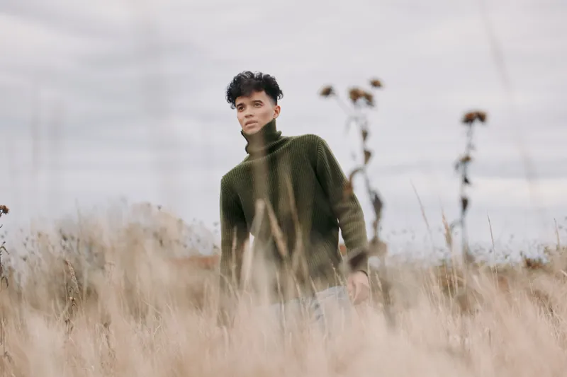 Young person standing in tall dry grass wearing a green sweater, captured in an authentic, natural pose that emphasizes genuine presence, quiet resilience and credible candid portrait storytelling.