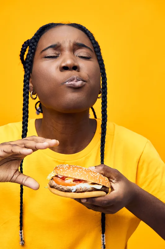Young woman enjoying a delicious hamburger with a vibrant yellow background, expressing happiness and satisfaction while indulging in fast food