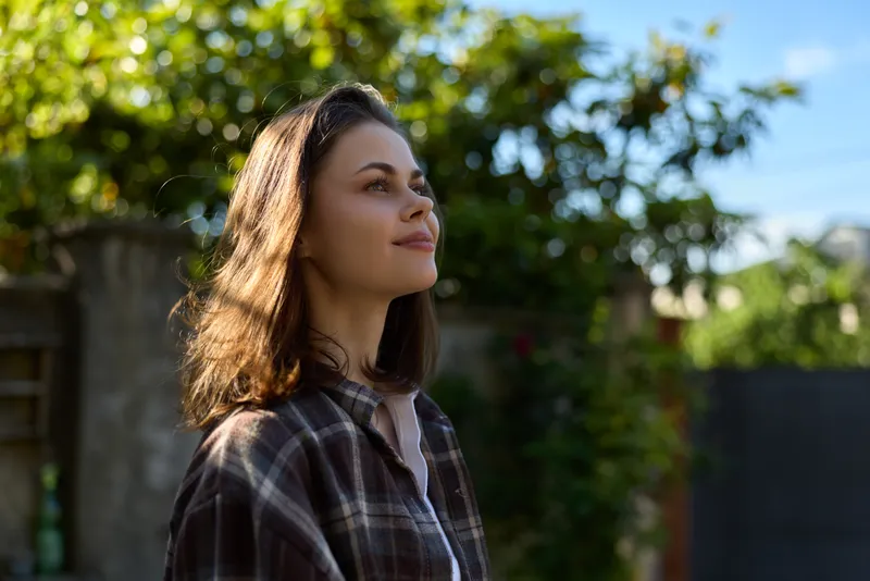 Woman outdoors, smiling softly, sunlight on her face, casual plaid shirt, relaxed pose, natural colors, lush greenery background, lifestyle moment, bright