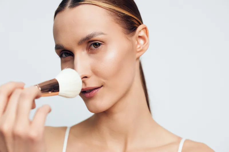 Young woman applies powder makeup on face using soft brush closeup portrait with natural skin tone and neutral background in beauty concept.