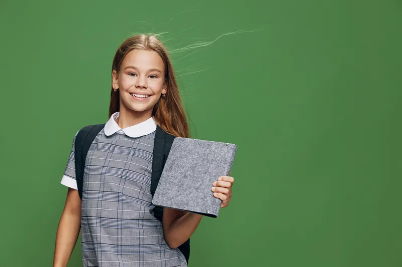 Smiling girl in school uniform holding tablet, standing with confidence on green background for education, technology, and digital learning concept.