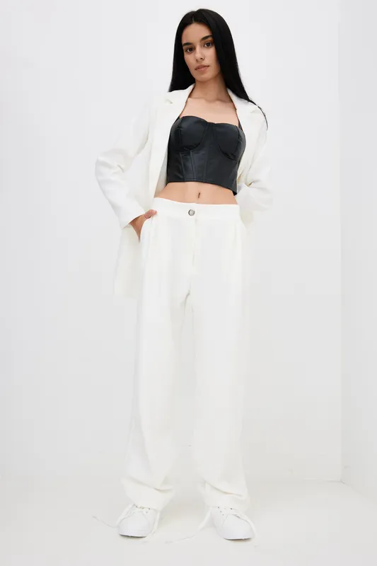 Stylish woman wearing a black bustier and white oversized pants with a matching white blazer, standing confidently against a minimalistic white background in modern fashion.