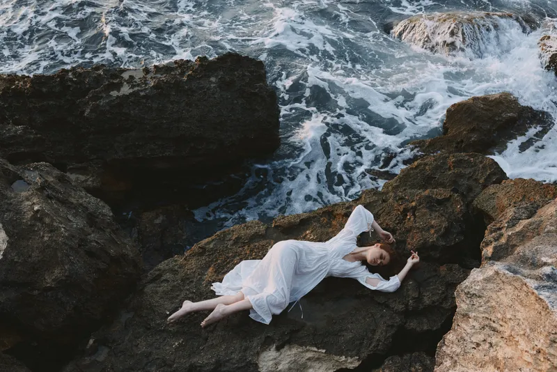 Красивая невеста в длинном белом платье с мокрыми волосами лежит на скалистом утесе