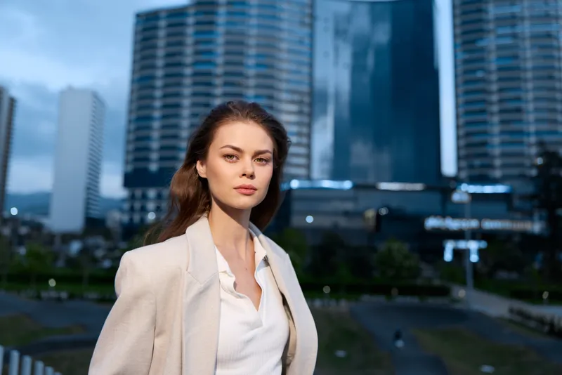 confident woman in business casual outfit standing outdoors with modern cityscape background at dusk, emotional and professional expression