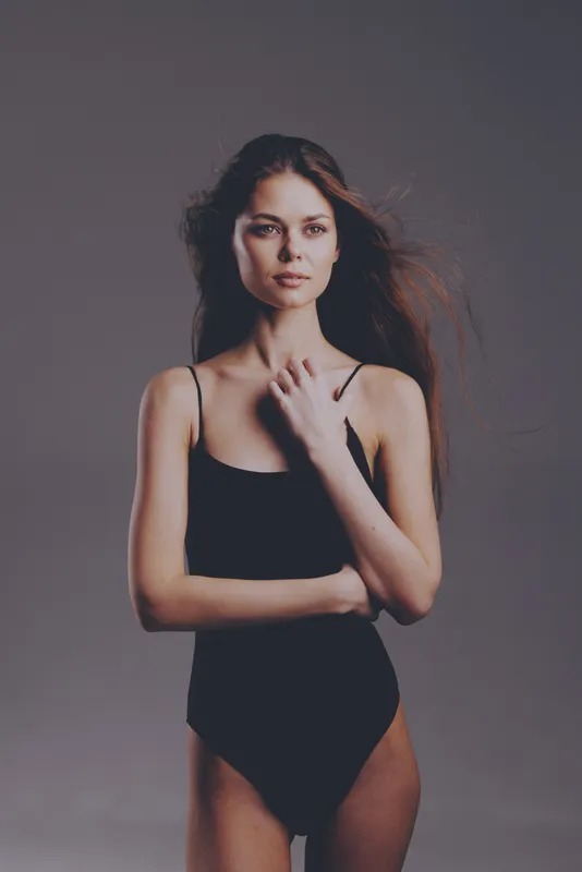 Young woman in black bodysuit posing confidently with flowing hair against neutral background