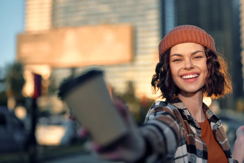 Woman smiling while taking a selfie with coffee in urban street during golden hour glow, candid lifestyle portrait conveying authenticity, mindful living and emotional storytelling in city.