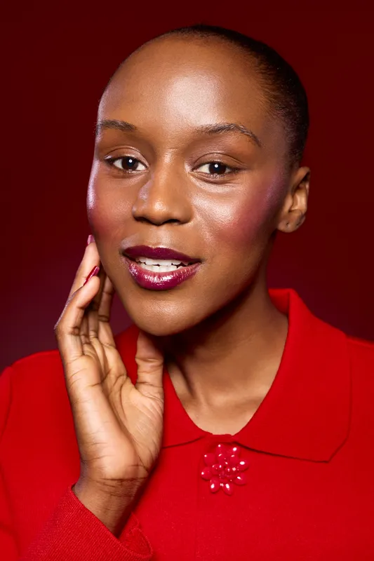 Close-up portrait of confident dark skinned African woman wearing red blouse, touching face with hand, smiling gently. Studio lighting highlights natural beauty and elegant makeup on burgundy