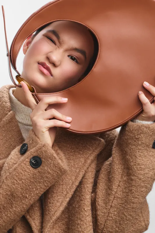 Asian woman is posing with a round brown bag in a warm, stylish studio. She wears a cozy coat and gazes softly at the camera, conveying confidence and calm in a contemporary fashion mood.