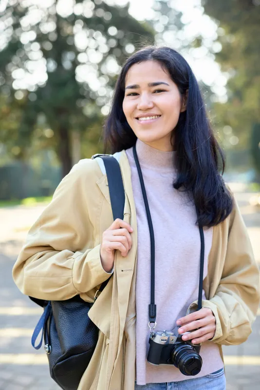 Smiling woman with a camera hanging from her neck and a backpack outdoors during daytime, casual jacket ready for travel and street photography shoot