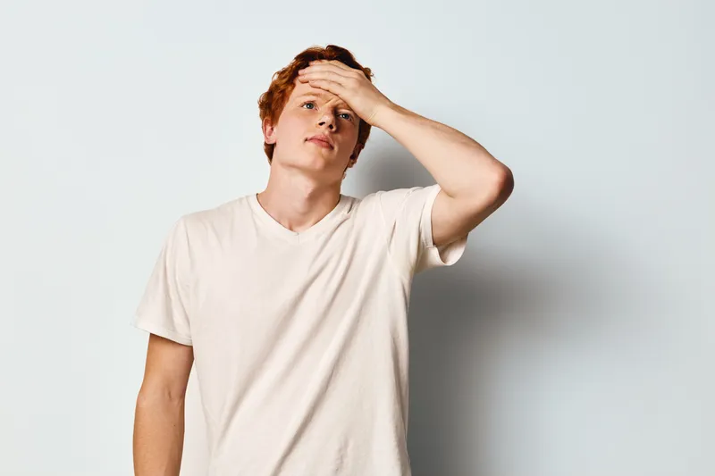 Young man with red hair in white t-shirt stands with hand on forehead looking thoughtful and concerned against white background, expressing stress or fatigue.