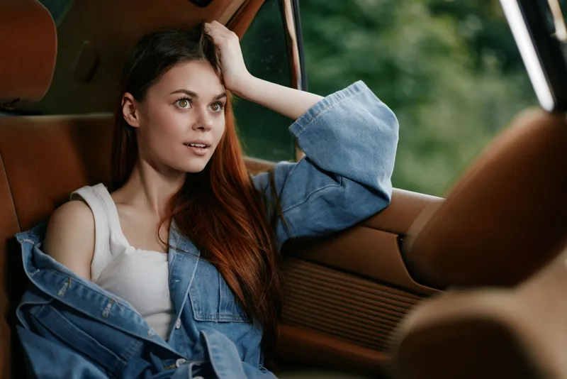 Young woman with long hair in denim jacket, sitting in a car, looking thoughtful and relaxed, with green nature background, summer vibes and casual style.