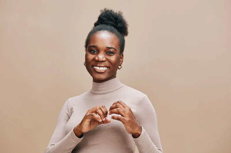 Cheerful Black woman in a beige turtleneck making a heart gesture, expressing love and positivity against a warm neutral background, ideal for health and wellness themes