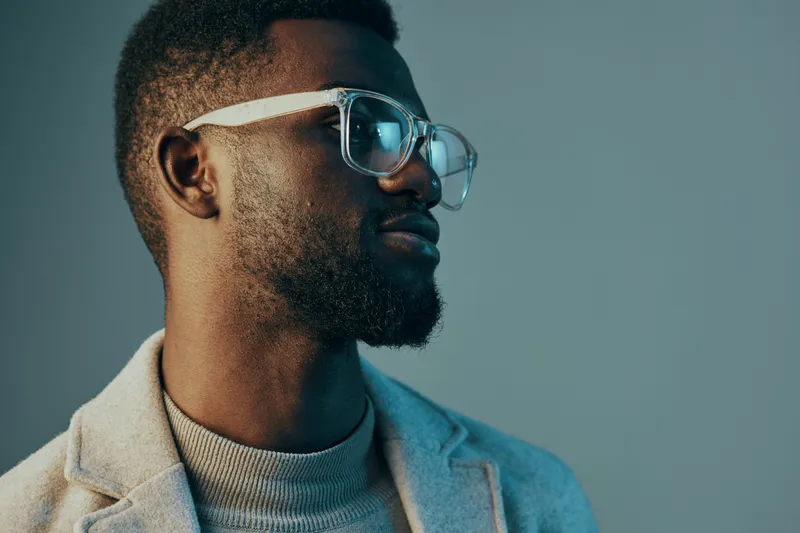 Confident young man with glasses in a stylish outfit, showcasing modern fashion and self assurance on a muted blue background