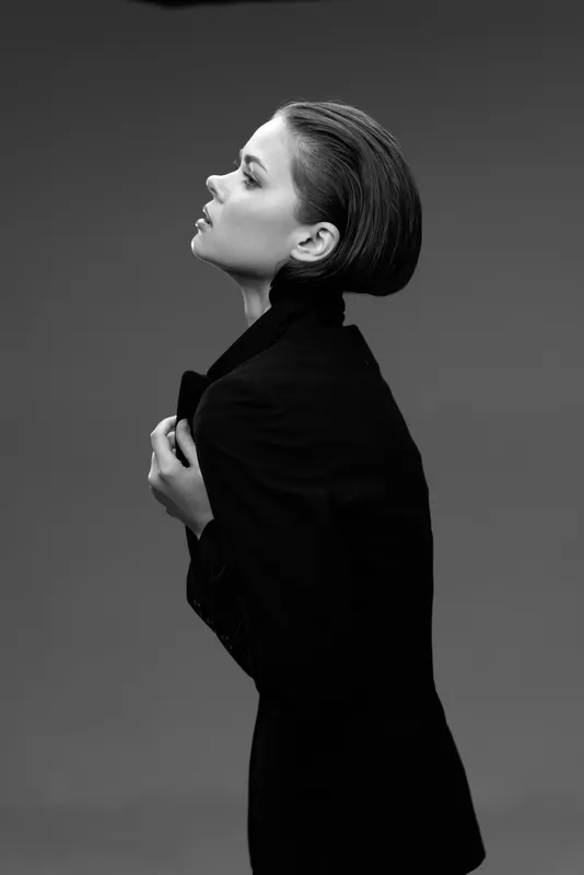 Black and white portrait of a stylish woman wearing a dark coat, posing with elegance and confidence in a studio setting.